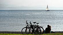 Bald mehr Ruhe am Bodensee? Ein Paar sitzt in Langenargen am Ufer, während im Hintergrund auf dem Bodensee ein Boot segelt.