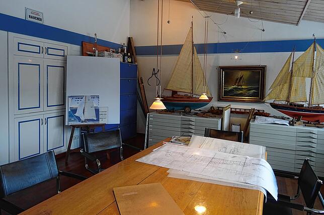 Das Büro von Josef Martin, maritim, mit Yachtmodellen. Rechts hängt eins von zwei Gemälden, das eine Yacht bei hohem Wellengang zeigt. ...