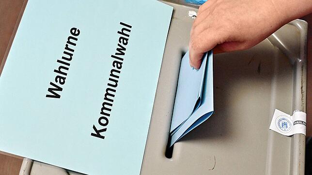 Eine Person steckt ihren Stimmzettel in eine Wahlurne mit der Aufschrift „Wahlurne Kommunalwahl“.