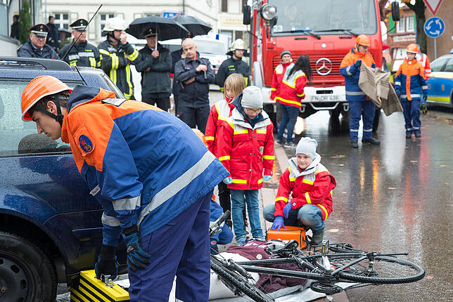 Die Jugendfeuerwehr hat das Fahrzeug angehoben und gesichert, die Kindergruppe des DRK-Ortsvereins kümmert sich um die Verletzte.