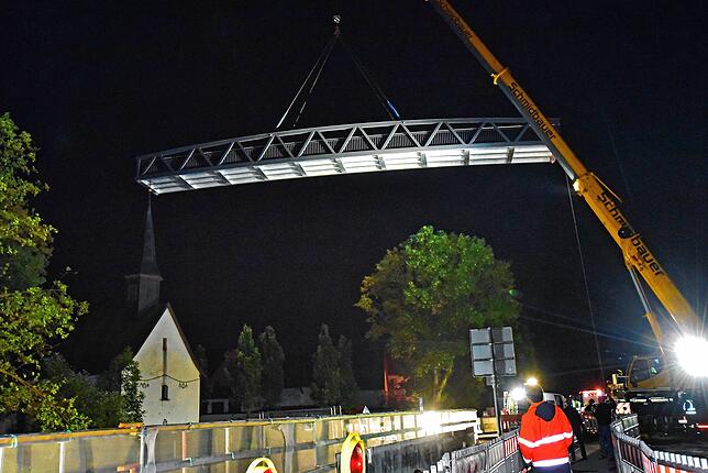 Die Alu-Brücke schwebt am Kranarm hängend über die Baustelle (im Hintergrund die Leonhardskapelle).