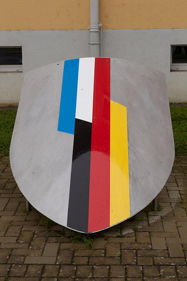 Vor der Kaserne ist ein Wappen in den Boden eingelassen, auf dem die Landesfarben beider Nationen ineinander übergehen.