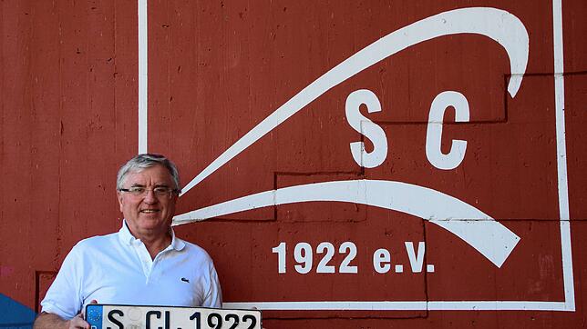 SC-Vorsitzender Thomas Kummer vor dem Logo seines Vereins. In der Hand hält er das Nummernschild eines Vereinskameraden, der zeitweise ...