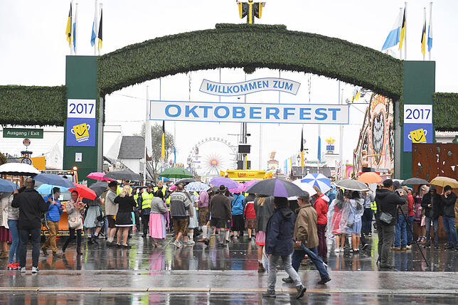 Vor dem Haupteingang stehen auf der Theresienwiesebeim Auftakt zum Oktoberfest Besucher mit Schirmen.