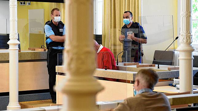 Der angeklagte Erpresser (im roten Pullover) sitzt zwischen Justizbeamten auf der Anklagebank und liest ein Dokument. Bild: dpa