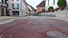 Die dunkelrote Fläche ist das neue, farbig asphaltierte Mobilitätsband in der Marktstraße. Im vorderen Bereich sind Abdeckungen und ...