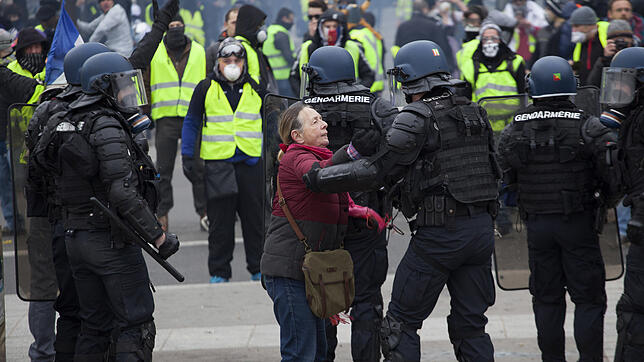 Vermummte Demonstranten stehen Polizisten gegenüber, von denen einer mit einer älteren Frau spricht.