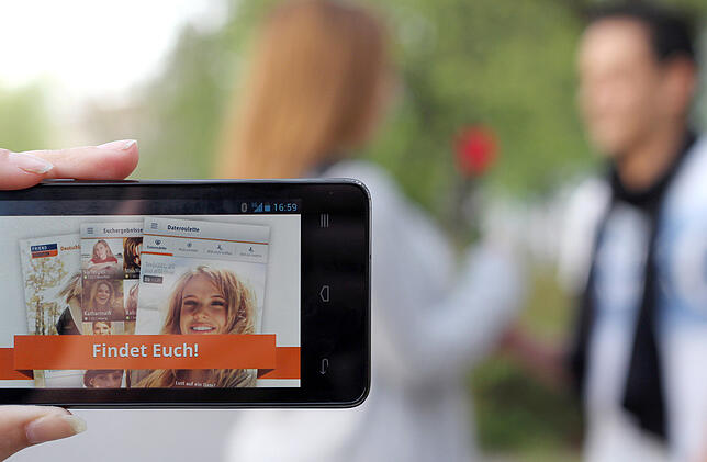Ein Mann und eine Frau reichen sich symbolisch eine Rose, während im Vordergrund die Internetseite eines Dating-Portals auf einem ...