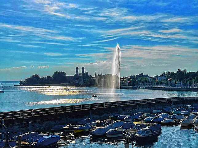 Die Fontäne in Friedrichshafen glitzert im Wasser vor der Silhouette der evangelischen Schlosskirche. Im Vordergrund der Gondelhafen.