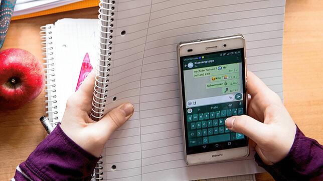 Eine Schülerin sitzt mit ihrem Handy und ihren Schulsachen an einem Schreibtisch. Soll die private Smartphone-Nutzung an Schulen ...